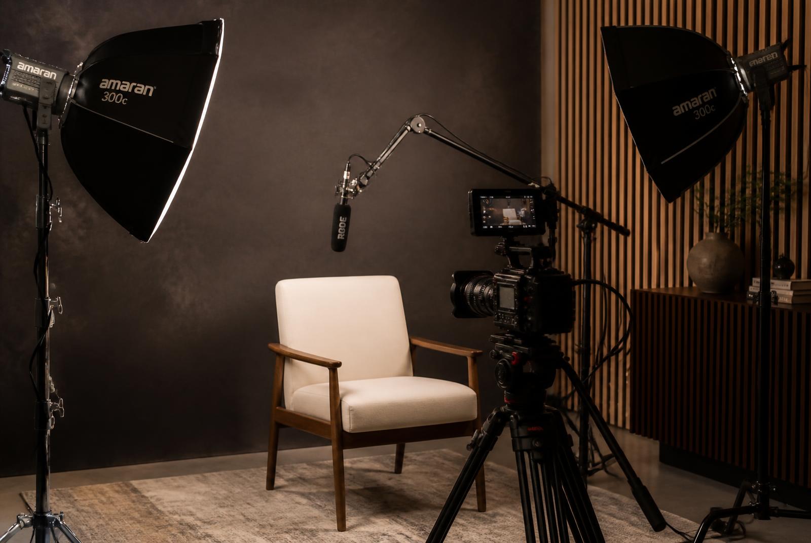 Filming studio with Amaran softbox lights, a cinema camera on a tripod, and a Rode microphone on a boom arm facing a subject chair